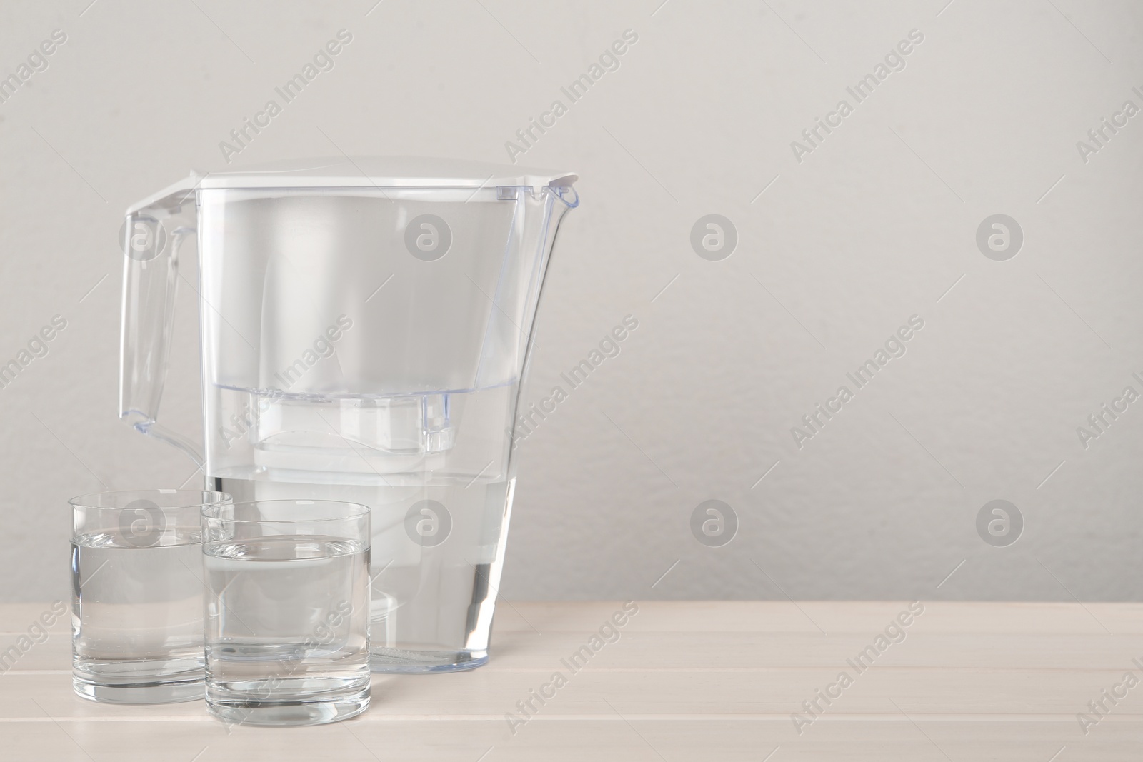 Photo of Filter jug and glasses with purified water on white table against light background. Space for text