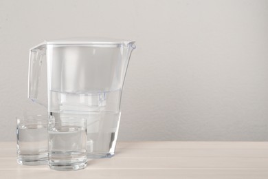 Photo of Filter jug and glasses with purified water on white table against light background. Space for text