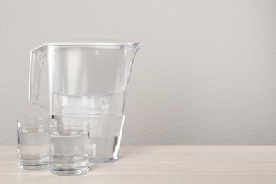 Filter jug and glasses with purified water on white table against light background. Space for text