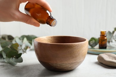 Photo of Woman dripping eucalyptus essential oil from bottle into bowl at light grey table, closeup