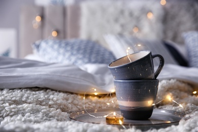 Photo of Cups, burning candle and garland on bed in room, space for text. Interior elements
