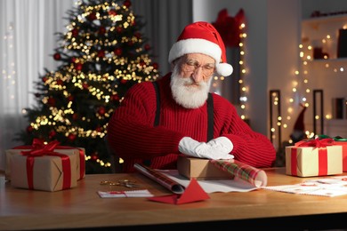 Photo of Santa Claus wrapping gift at his workplace in room decorated for Christmas