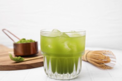 Photo of Glass of delicious iced green matcha tea, powder and bamboo whisk on white tiled table, closeup