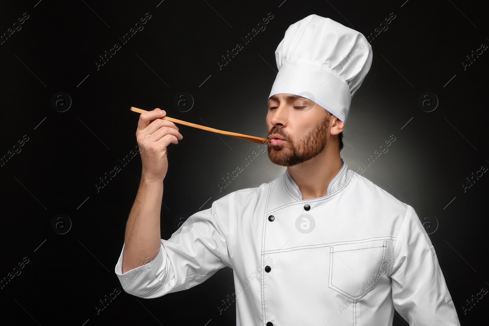 Photo of Mature chef with spoon tasting dish on black background