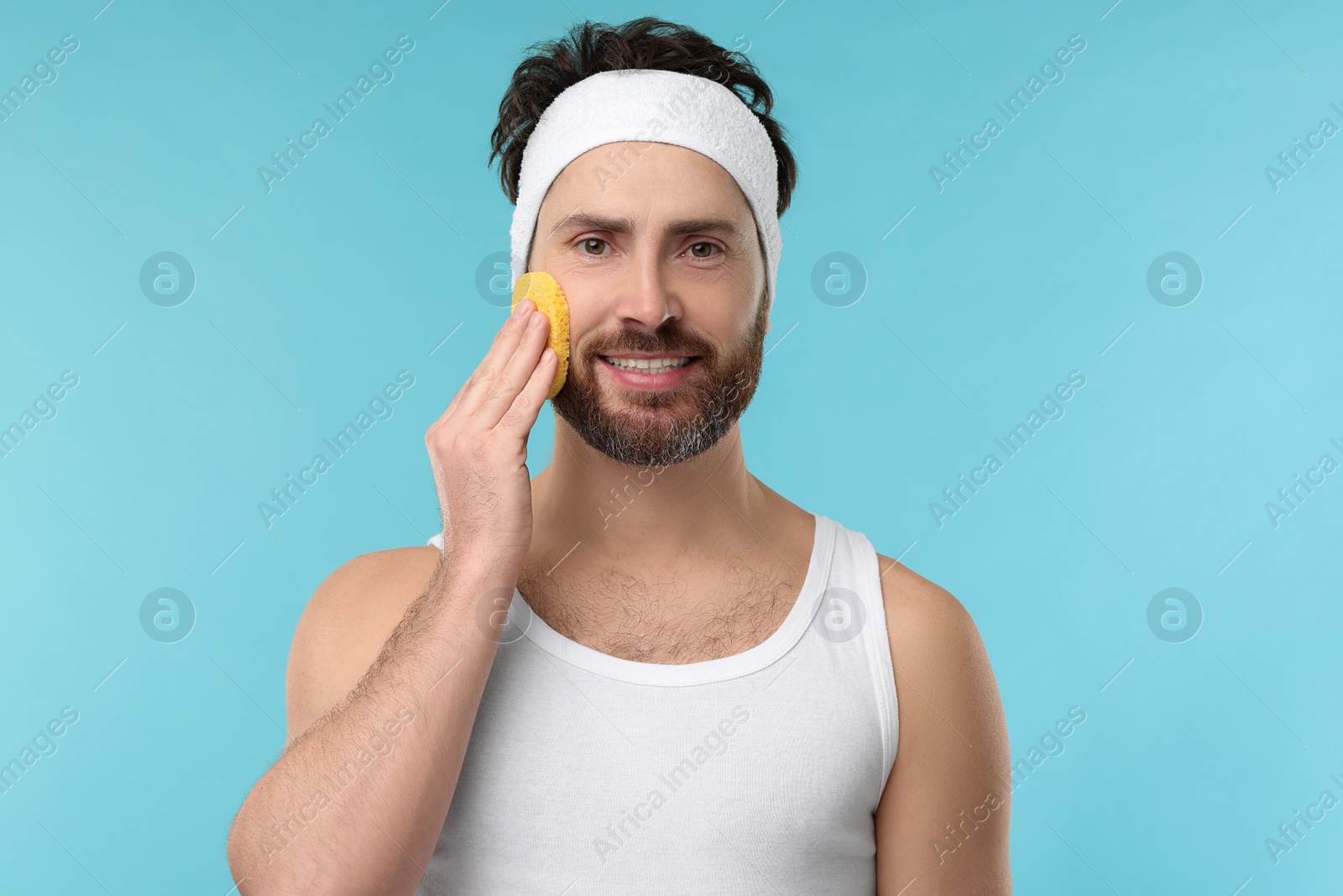 Photo of Man with headband washing his face using sponge on light blue background