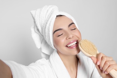 Woman in bathrobe with towel taking selfie while singing into brush on light grey background. Spa treatment