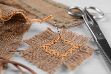 Photo of Pieces of burlap fabric with stitches, needle and scissors on white wooden table, closeup