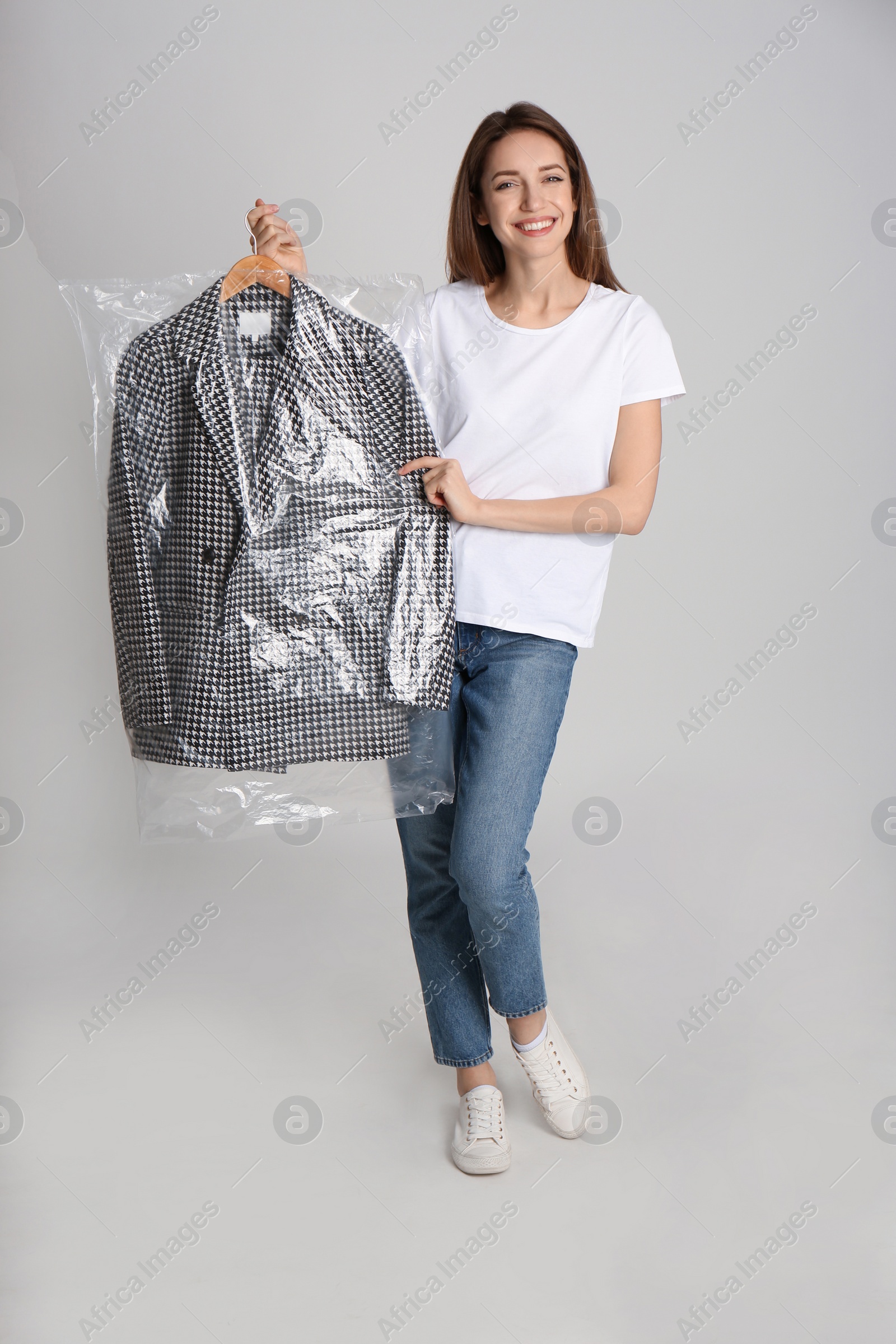 Photo of Young woman holding hanger with jacket in plastic bag on grey background. Dry-cleaning service