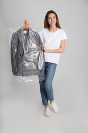 Young woman holding hanger with jacket in plastic bag on grey background. Dry-cleaning service