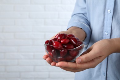 Woman with glass bowl of fresh ripe dogwood berries near white brick wall, closeup