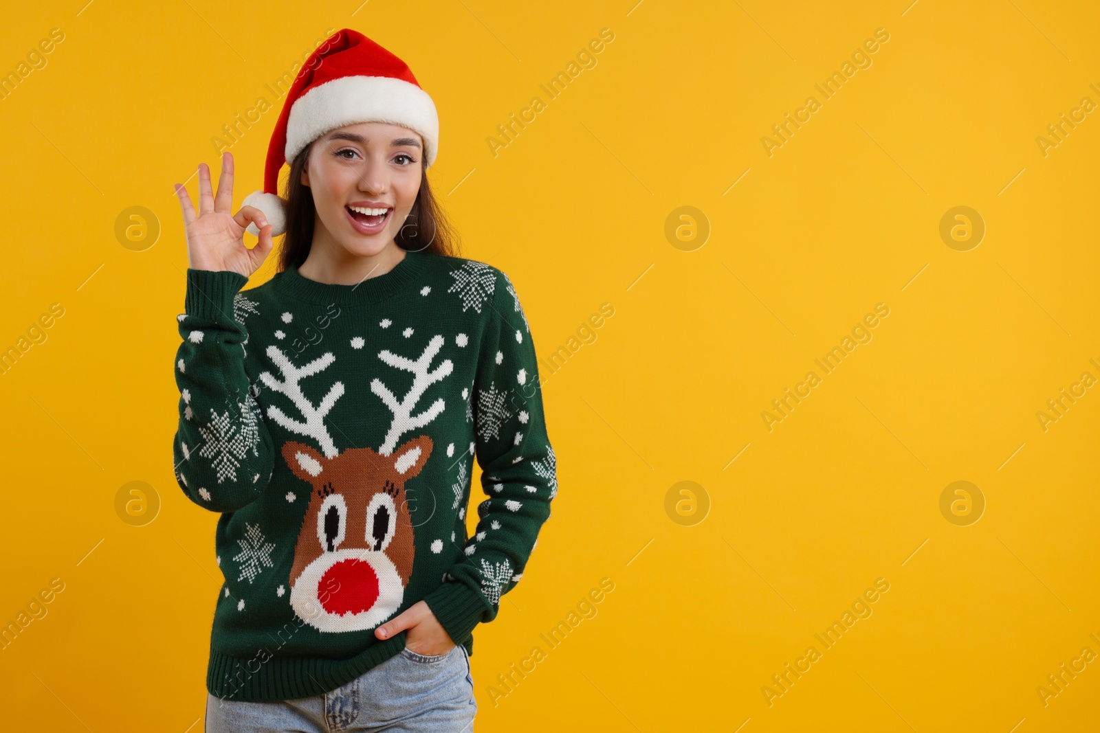 Photo of Happy young woman in Christmas sweater and Santa hat showing OK gesture on orange background. Space for text