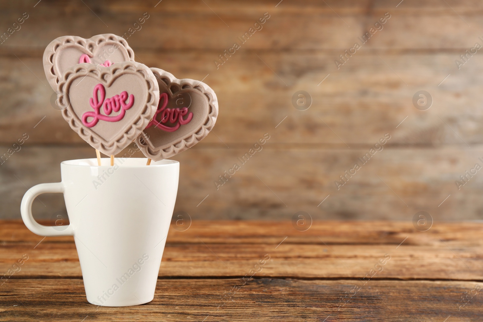 Photo of Heart shaped lollipops made of chocolate in cup on wooden table, space for text