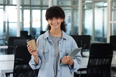 Happy woman with paper cup of drink and tablet in office