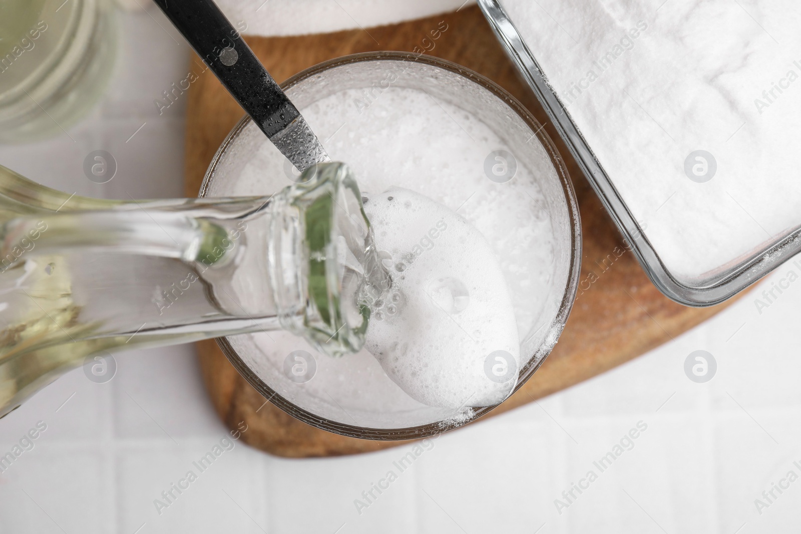 Photo of Pouring vinegar into spoon with baking soda over bowl at white tiled table, top view