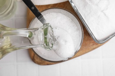 Pouring vinegar into spoon with baking soda over bowl at white tiled table, top view