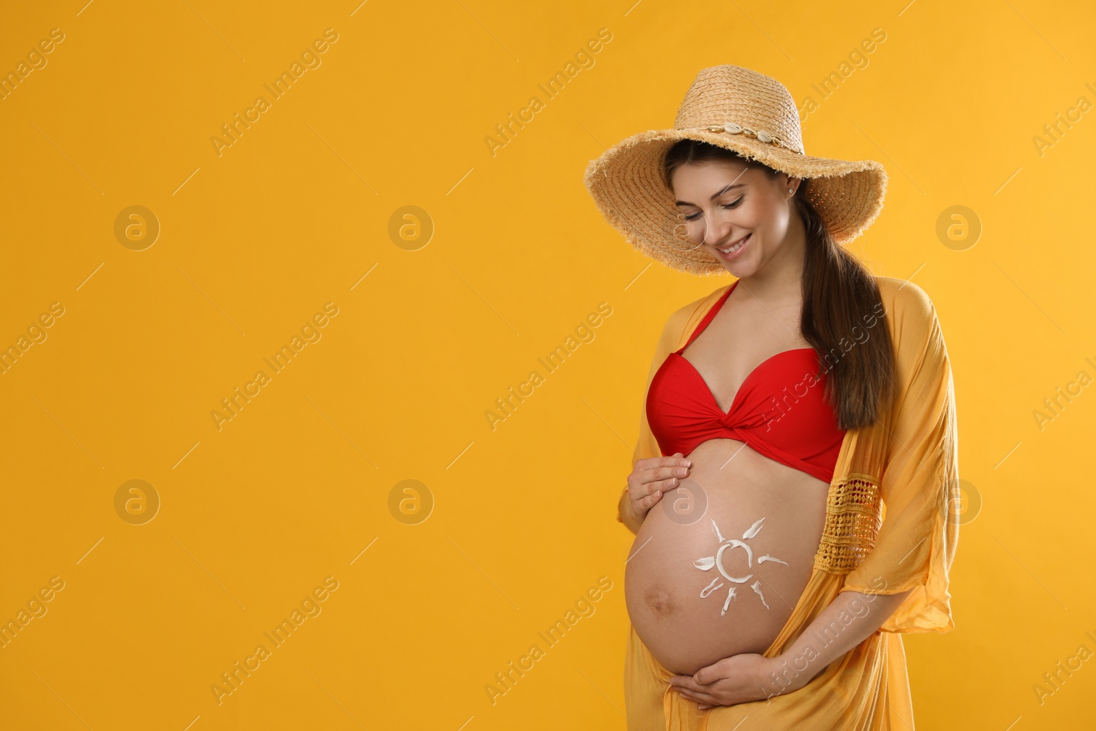 Photo of Young pregnant woman with sun protection cream on belly against yellow background, space for text