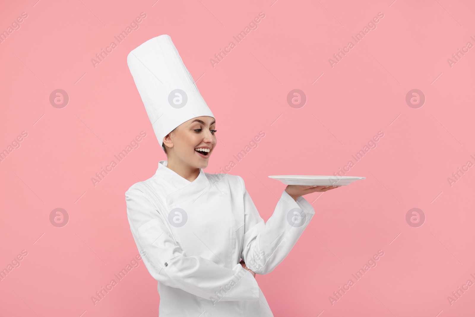 Photo of Happy professional confectioner in uniform holding empty plate on pink background