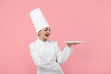 Happy professional confectioner in uniform holding empty plate on pink background