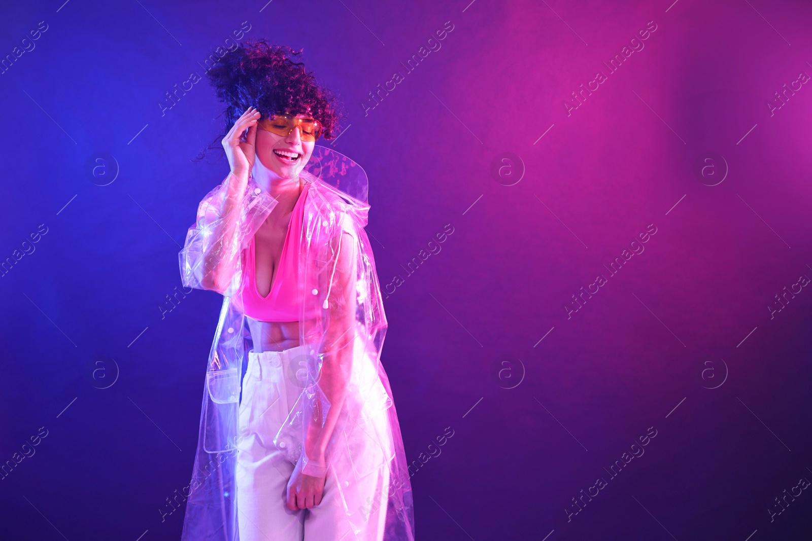 Photo of Beautiful young woman in transparent coat and sunglasses posing on color background in neon lights. Space for text