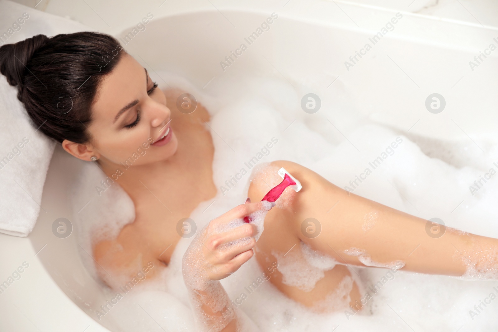 Photo of Young woman shaving legs in bubble bath, above view