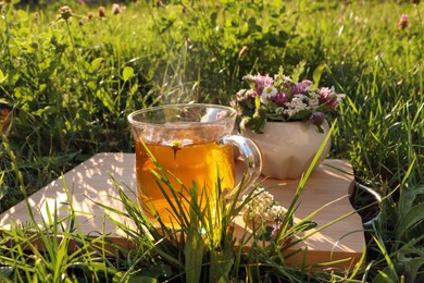 Photo of Cup of aromatic herbal tea and ceramic mortar with different wildflowers on wooden board in meadow