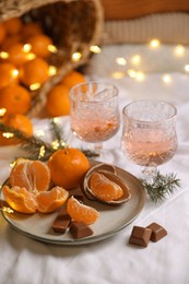 Photo of Delicious ripe tangerines, chocolates, festive lights and glasses of wine on white bedsheet