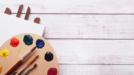 Photo of Artist's palette with samples of colorful paints, brushes and canvas on white wooden table, flat lay. Space for text