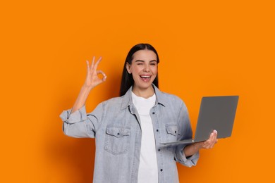Photo of Happy woman with laptop winking and showing okay gesture on orange background