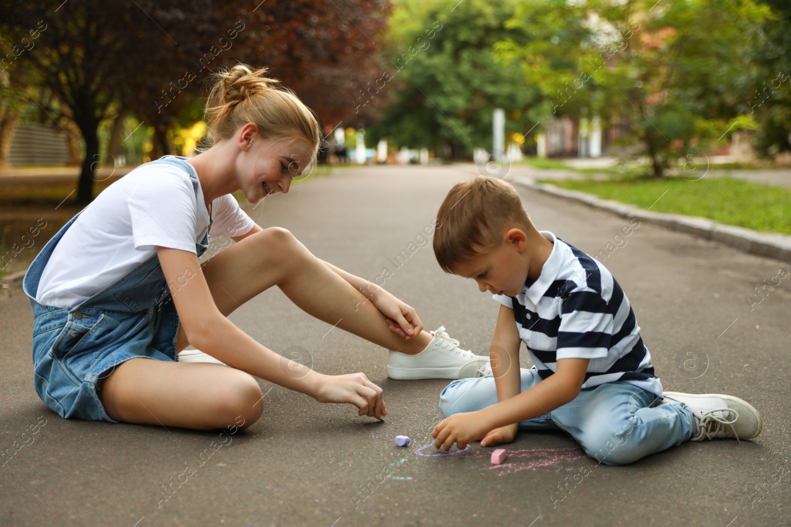 Photo of Teen nanny and cute little boy drawing with chalks on asphalt