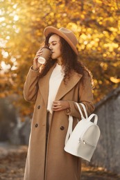 Beautiful African-American woman with stylish white backpack drinking coffee on city street