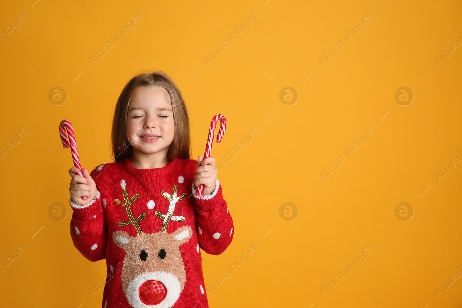 Photo of Cute little girl in Christmas sweater holding candy canes on yellow background, space for text