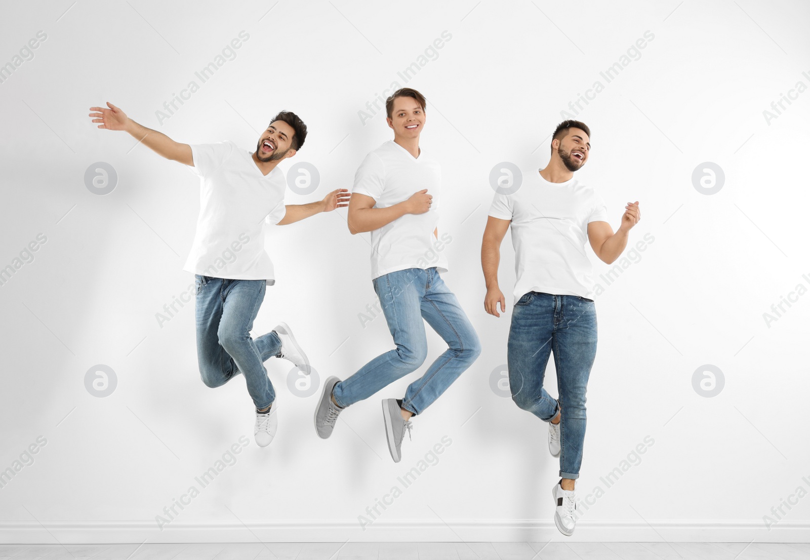 Photo of Group of young men in stylish jeans jumping near white wall