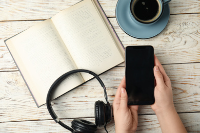 Woman holding mobile phone over white wooden table with book, top view