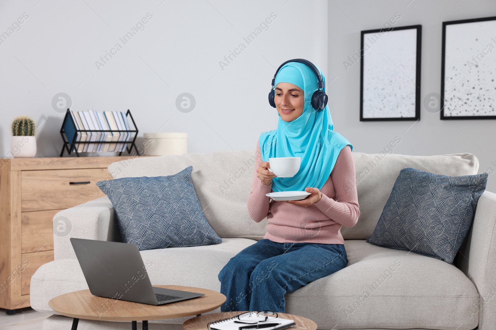 Photo of Muslim woman with cup of drink using laptop at wooden table in room