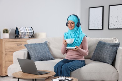 Muslim woman with cup of drink using laptop at wooden table in room