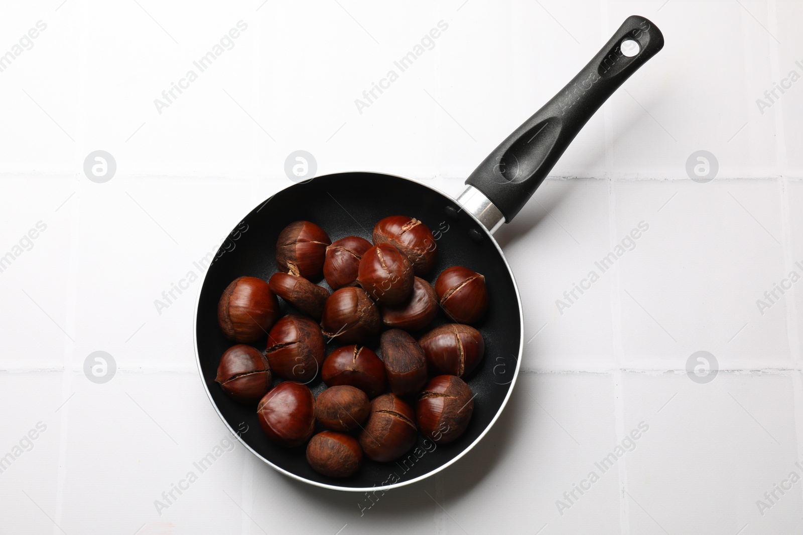 Photo of Fresh edible sweet chestnuts in frying pan on white tiled table, top view