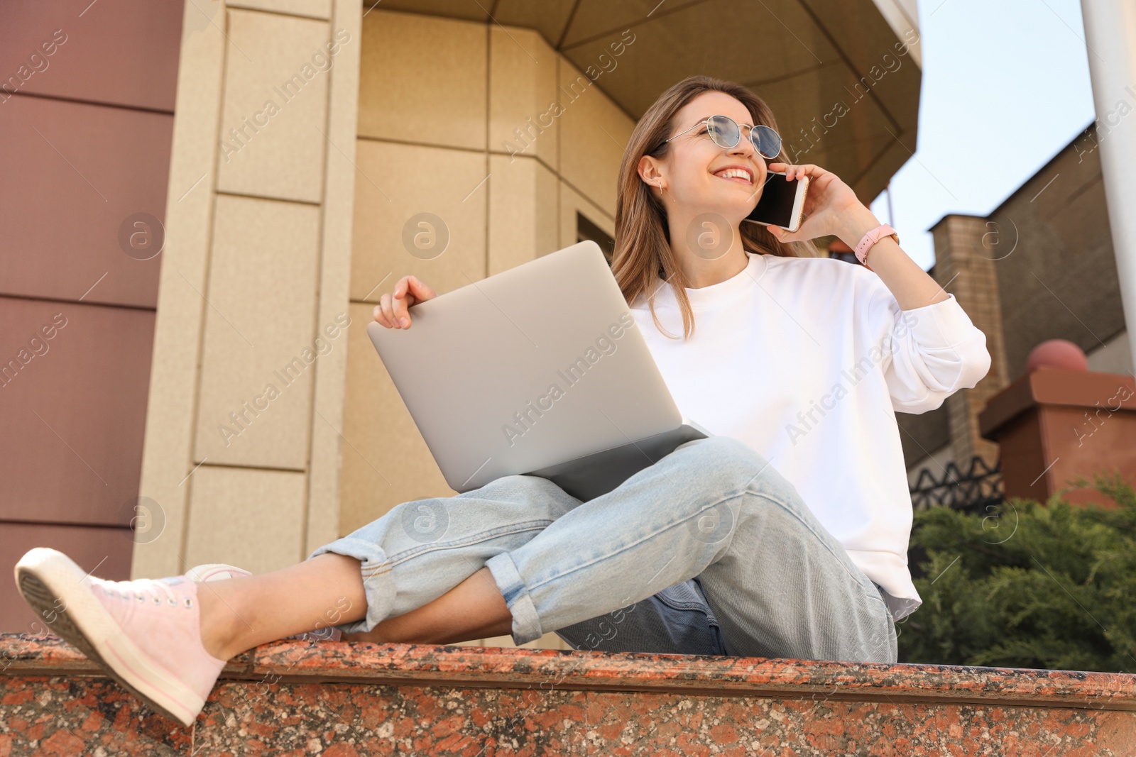 Image of Happy young woman with laptop talking on phone outdoors