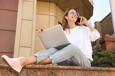 Happy young woman with laptop talking on phone outdoors