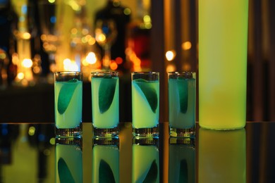 Photo of Shot glasses with alcohol drink and lime wedges on mirror surface against blurred background