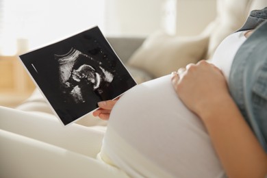 Pregnant woman holding ultrasound picture near her belly indoors, closeup