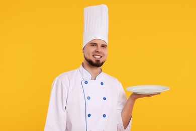 Photo of Happy professional confectioner in uniform holding empty plate on yellow background