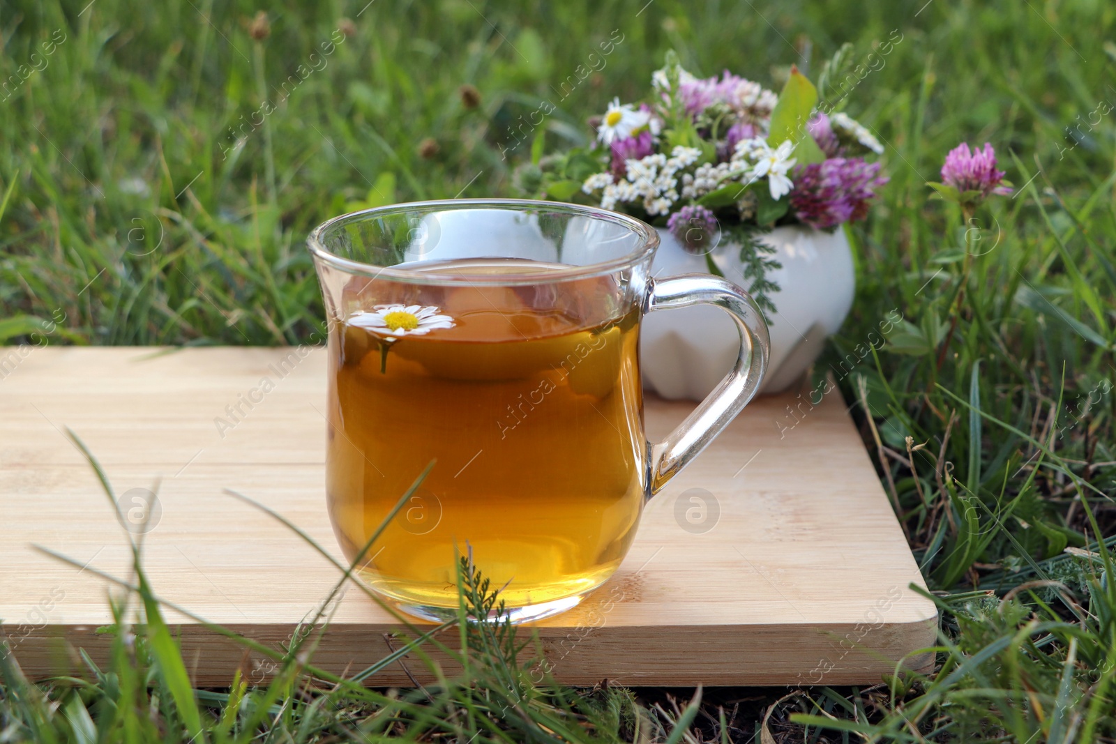 Photo of Cup of aromatic herbal tea, pestle and ceramic mortar with different wildflowers on green grass outdoors