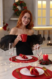 Photo of Beautiful young woman with cup of drink and smartphone at table served for Christmas in kitchen