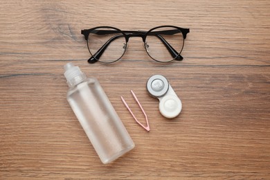 Case with contact lenses, tweezers, glasses and drops on wooden table, flat lay