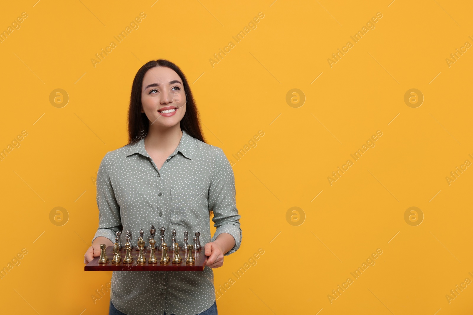 Photo of Happy woman holding chessboard with game pieces on orange background, space for text