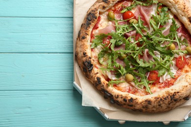 Photo of Tasty pizza with meat and arugula in cardboard box on light blue wooden table, above view. Space for text