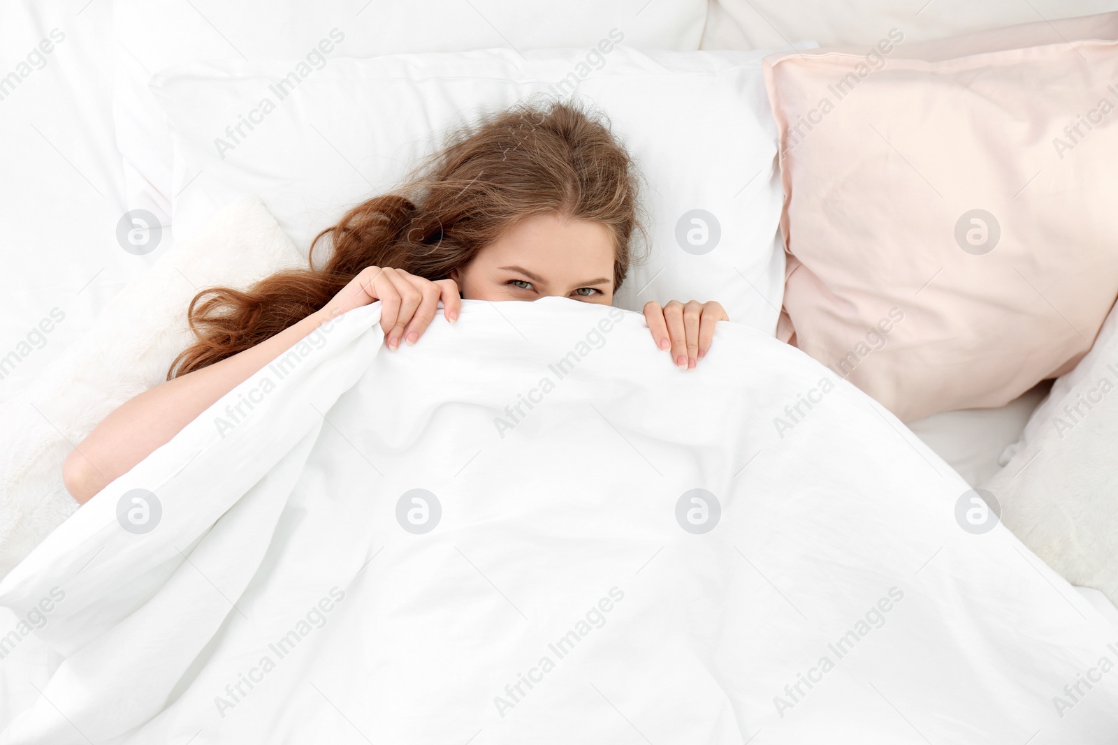 Photo of Young woman lying on soft pillows and hiding under blanket in bed at home