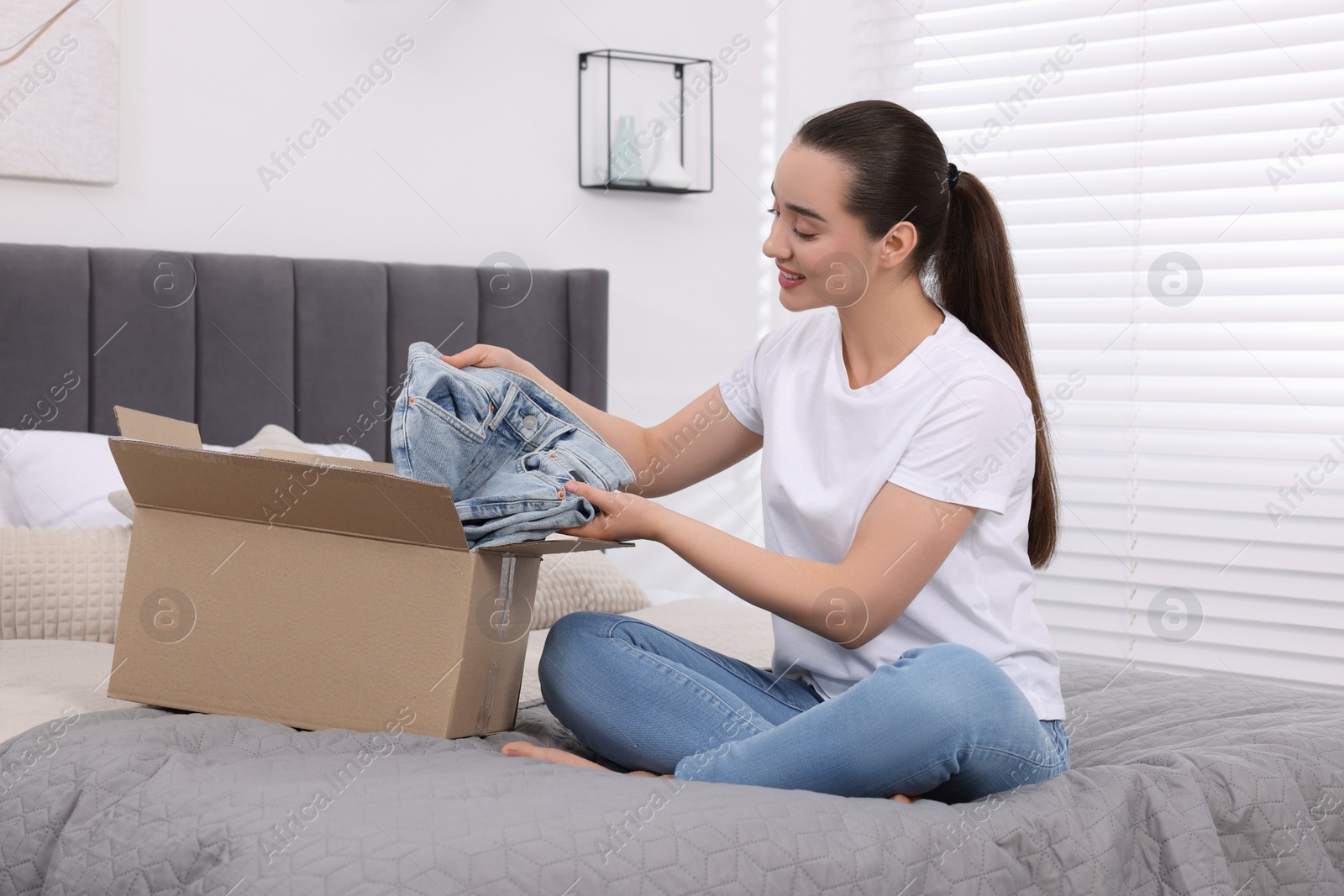 Photo of Happy young woman taking jeans out of box on bed at home. Online shopping