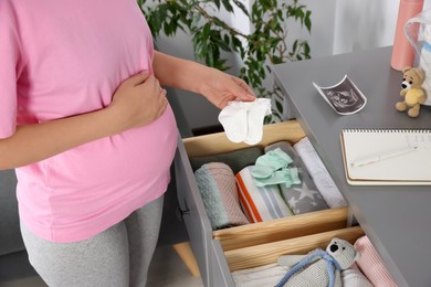 Photo of Pregnant woman taking cute baby socks from open drawer at home, closeup