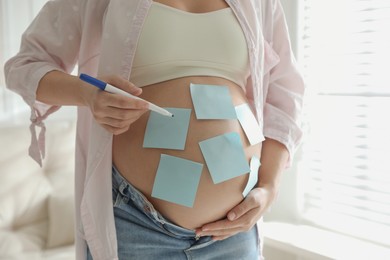 Photo of Pregnant woman with marker and sticky notes on belly indoors, closeup. Choosing baby name